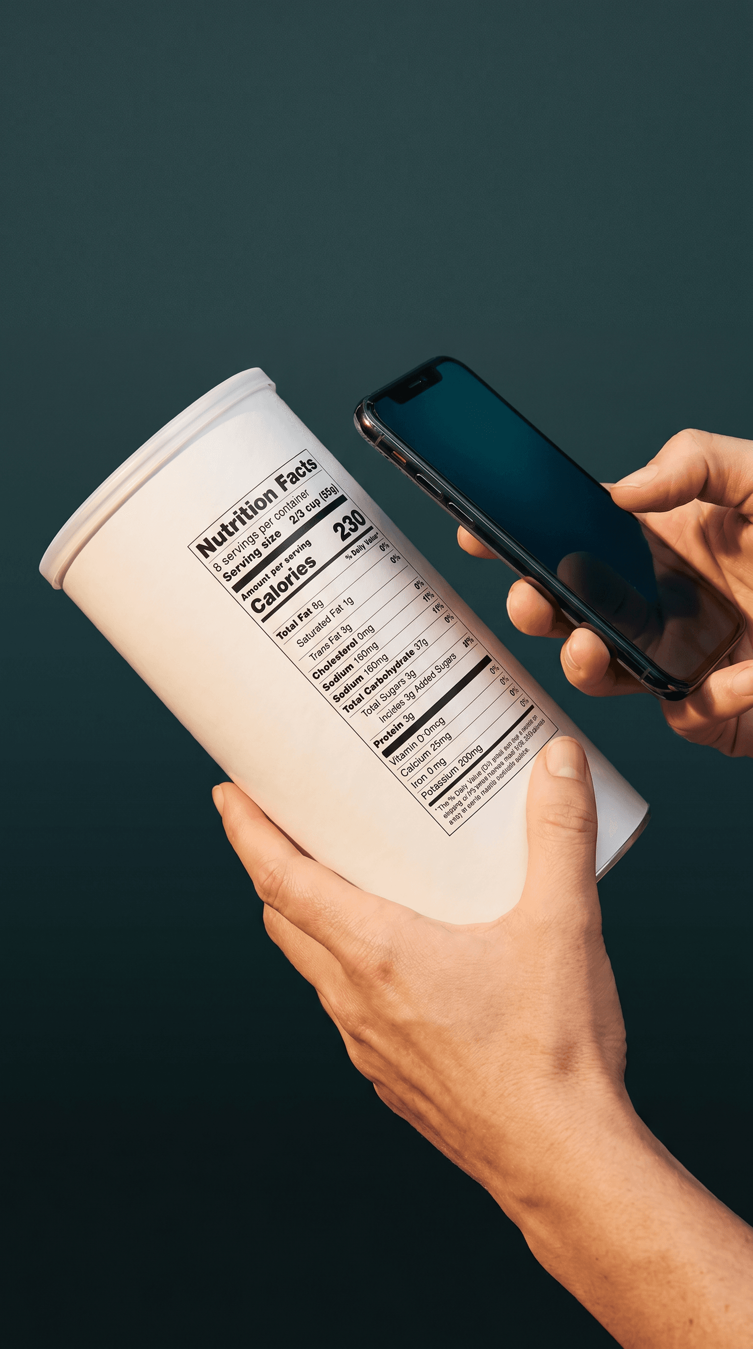 Editorial close-up photograph of hands holding a product canister with a clearly-visible Nutrition Facts label, an iPhone hovering parallel to the label in the act of scanning, on a dark teal background
